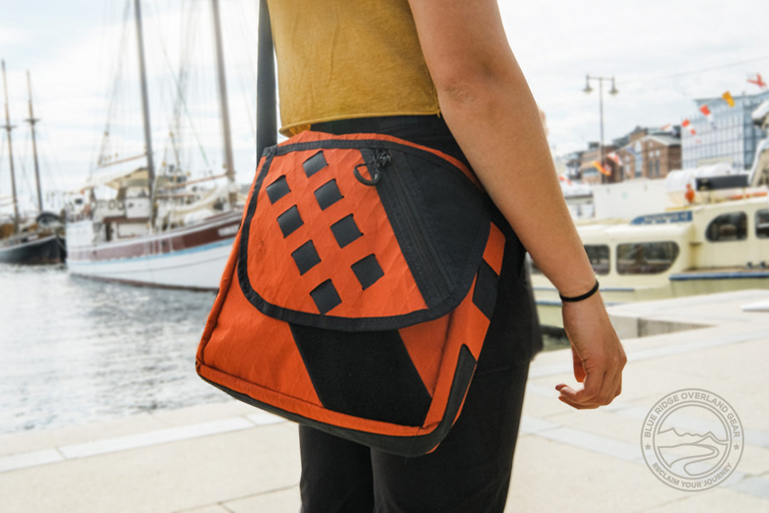 Orange Transit Bag being carried by a young woman who is traveling the world, near the edge of a dock with sail boats